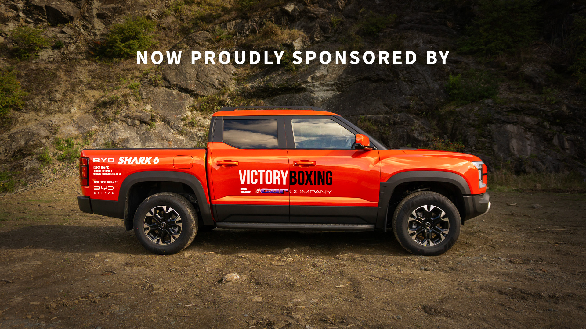 BYD Shark 6 in orange, parked in front of rocks with Victory Boxing signage on it.